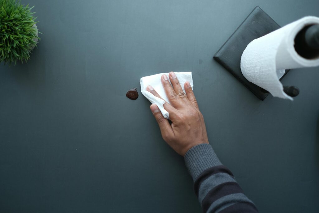 A hand wipes a dark surface with a tissue near a paper roll and plant.